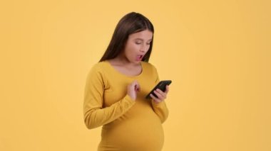 Pregnancy shopping. Young pretty pregnant woman web surfing on phone, choosing cute baby stuff, touching her belly, orange studio background, slow motion, free space