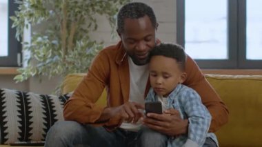 Kids and gadgets in everyday life. Lovely little black boy cuddling with daddy and watching his father playing online games on smartphone, resting at home, tracking shot, empty space