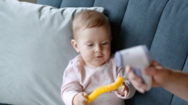 Health check. Close up portrait of adorable little baby girl playing with toy on sofa, dad hand measuring body temperature with contactless thermometer at home, slow motion, empty space