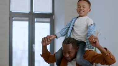 Fun with daddy. Loving happy african american father riding his cute little son on shoulders, boy laughing, enjoying game at home, tracking shot, slow motion, empty space