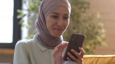Domestic leisure. Young happy muslim lady wearing traditional headscarf reading news on smartphone online, web surfing in social media, resting on weekend at home, tracking shot, free space