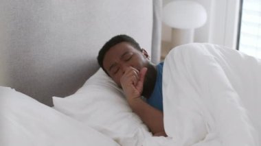 Sickness symptoms. Young black african american man sleeping in bed but waking up with cough attack, keep resting at home in morning, tracking shot, slow motion, free space