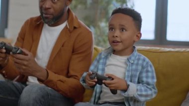 Father and son entertainment. Cute little african american boy playing video games with joystick at home, competing with his emotional daddy in car race, tracking shot, slow motion, free space