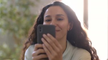 Close up portrait of young pretty middle eastern woman reading news in social media, resting at home in morning, sun flare, slow motion, tracking shot