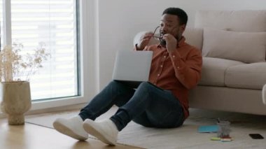 Overwork concept. Young african american man working on laptop, getting tired, taking off his eyeglasses and rubbing nose bridge, sitting on floor at home, tracking shot, slow motion, empty space