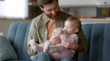 Baby care. Loving young father holding his little daughter on hands, adorable thirsty girl drinking water from bottle, resting together at home, tracking shot, slow motion, free space