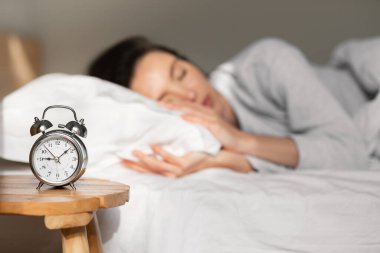 Tired young european lady lies and sleeps on white comfortable bed, selective focus on alarm clock on table in bedroom interior, blurred. Time to wake up, overslept, late for work, meeting at home