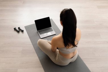 High angle view of long-haired brunette athletic young woman in beige sportswear sitting on yoga mat at home, using modern laptop with black empty screen, mockup. Online fitness class concept