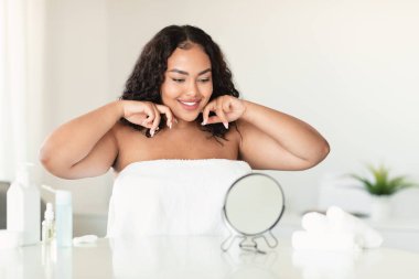 Beautiful body positive lady making lymphatic drainage facial self massage, looking at mirror, sitting in bedroom, free space. Plus size woman doing domestic skin care treatmenrt