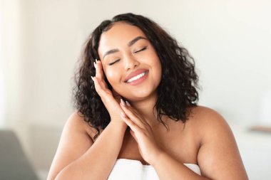 Beautiful happy overweight lady touching her face and smiling with closed eyes, positive bodypositive woman standing wrapped in towel in bathroom interior