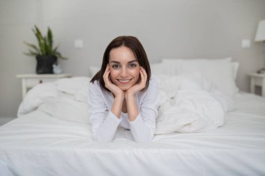 Good morning. Relaxed beautiful young brunette woman posing in bed at home, happy cheerful lady lying under blanket on her belly, smiling at camera, cozy bedroom background, copy space