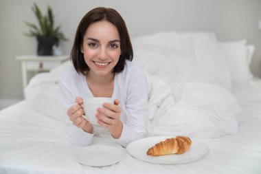 Portrait of cheerful happy beautiful brunette young woman in white pajamas have breakfast and smiling, lady drinking coffee, eating croissant in bed after waking up in the morning, copy space