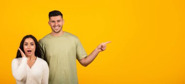 Cheerful shocked young arabic couple in casual with open mouth point finger at empty space isolated on yellow background, studio, panorama. Advice, reaction to good news, surprise, sale, ad and offer