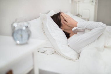 Insomnia, sleep disorder concept. Exhausted brunette young woman lying in bed at home, touching her face, feeling tired in the morning after sleepless night, alarm on bedside table, side view