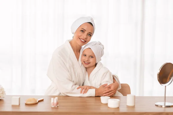 Beauty Portrait Of Happy Young Mom And Daughter In Bathrobes And Towels On Head Embracing At Home, Cute Little Girl And Her Mother Sitting At Table With Cosmetics, Having Spa Day Together, Copy Space