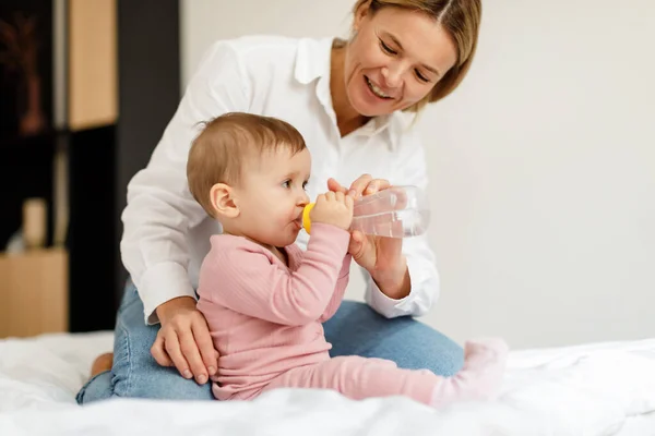 Baby hydration. Caring mother giving water bottle to her infant child, sitting on bed at home, copy space. Cute toddler girl enjoying healthy drink, spending time with mom