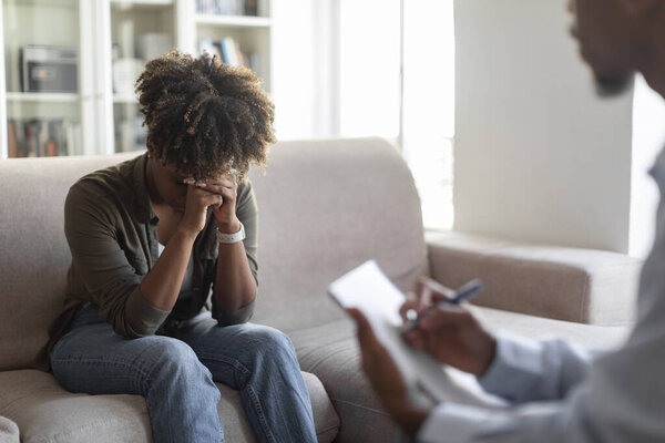 Upset young black woman in casual sitting on couch, cover face with palms, crying while have therapy session with man therapist, african lady fighting addiction, visiting psychologist, copy space