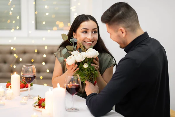 Happy attractive brunette young woman in nice outfit holding bouguet flowers and looking at her handsome boyfriend, happy loving couple have valentine celebration at home, copy space