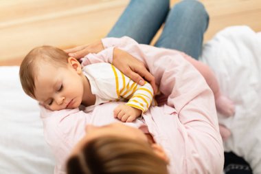 Caring mother holding her adorable small sleeping baby girl on hands, singing lullaby, comforting infant kid, sitting on bed in bedroom at home, above view