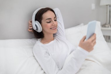 Cheery pretty young woman in white cotton pajamas chilling in bed with smartphone and wireless headphones, looking at gadget screen and smiling, watching movie or TV show on Internet, copy space