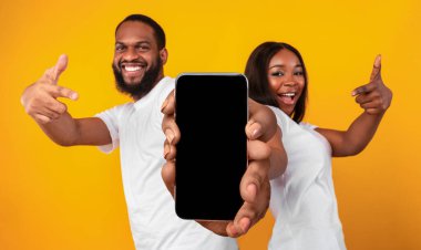 Check This. Black couple pointing at big blank smartphone screen for mockup, excited african american spouses recommending new mobile application or website while standing on yellow background
