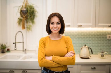 Candid photo of cheerful happy pretty brunette young lady in casual outfit posing in modern cozy kitchen, arms crossed on chest, smiling at camera, copy space. Millennials lifestyle concept