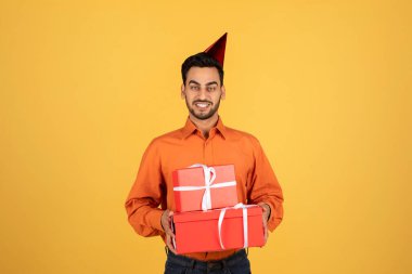 Handsome arab man holding wrapped gift boxes and wearing birthday hat while standing on yellow studio background, happy middle eastern male having B-day party, carrying presents, copy space