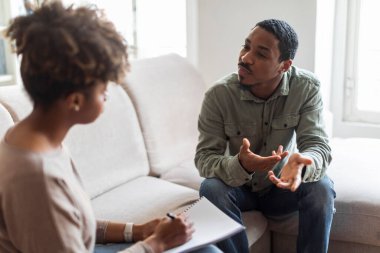 Psychological help concept. Confused handsome young black guy sitting on couch against african american woman therapist, gesturing and talking, patient sharing thoughts and feelings with psychologist