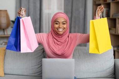 Excited black lady in hijab sitting on sofa with laptop and lifting gift bags above her head at home. Charming african american muslim woman purchasing presents on web, happy to get big discount