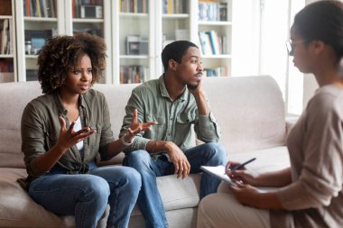 Attractive curly young black woman in casual sharing thoughts with african american lady psychologist during family session, frustrated spouse millennial man sitting be her, looking at copy space