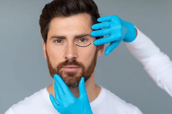 Portrait of middle aged man getting spa treatment from beautician in protective gloves, plastic surgeon making marks on male face, grey studio background