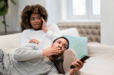 Scared shocked millennial african american man and woman couple spouses sitting on couch, gesturing, watching appalling content on TV, copy space for advertisement