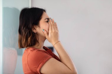 Mental Breakdown. Closeup Portrait Of Young Arab Woman Crying At Home, Upset Middle Eastern Female Leaning On Wall And Covering Face With Hands, Suffering Depression, Side View With Copy Space