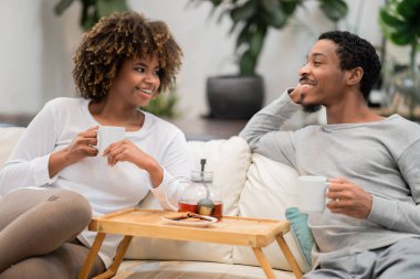 Positive cheerful relaxed millennial black lovers enjoying tea at home, happy african american man and woman in pajamas sitting on couch, chatting, resting at weekend together