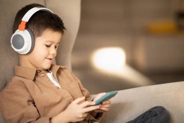 Smiling caucasian little kid in casual, wireless headphones watch video on phone sits in armchair in living room interior, close up. Online call, learning app, lifestyle and gadget addiction at home