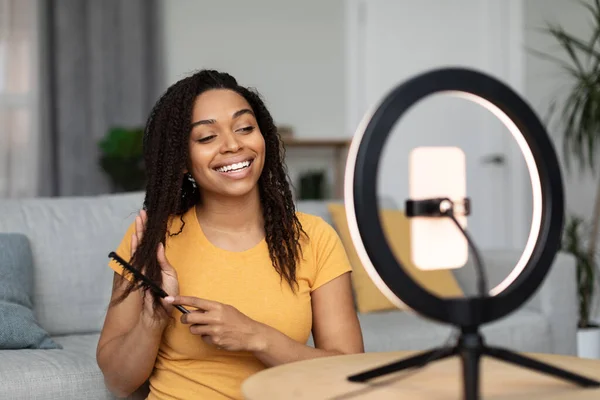Beauty blog concept. Young african american woman combing her hair and recording video on cellphone, talking about hair care. Lady talking at phone camera on ring lamp tripod
