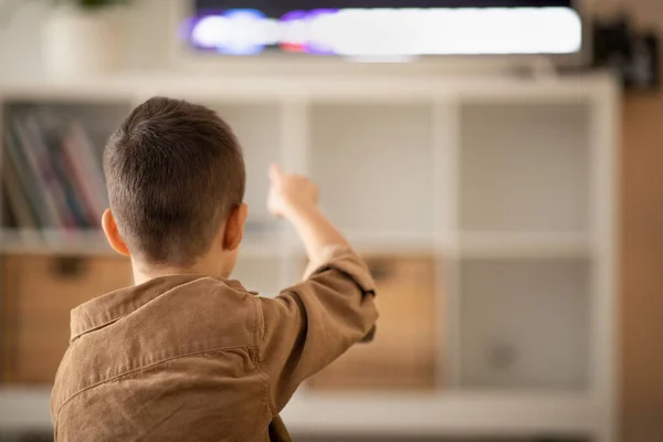 European small kid watch video, pointing finger at tv set, sits on floor in living room interior, back, close up, empty space. Education with device at home, cartoons at free time and video tutorials