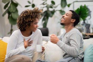 Happily married young african american couple in homewear sitting on couch in living room, holding coffee mugs, have conversation and laughing, family drinking coffee at home
