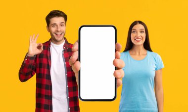 Cheerful Young Couple Holding Big Blank Smartphone In Hands And Showing Ok Gesture, Positive Man And Woman Advertising Online Offer Or Mobile Application While Standing On Yellow Background, Mockup