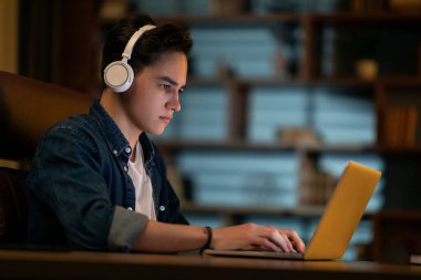 Concentrated young man IT specialist web-developer working on laptop alone at dark office, using wireless headphones, programmer listening to music while coding, side view, copy space