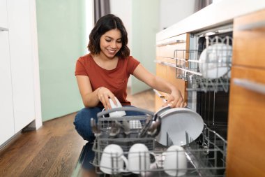 Portrait Of Happy Young Middle Eastern Woman Unloading Dishwasher Machine In Kitchen At Home, Smiling Arab Female Taking Out Clean Dry Cups And Dishes, Enjoying Making Domestic Chores, Free Space