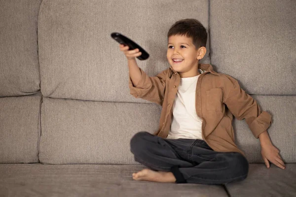 Happy small european boy with remote control watch video, has fun and shoots, switches channels on TV, sits on sofa at home, free space. Entertainment alone, game, childhood and cartoons at free time