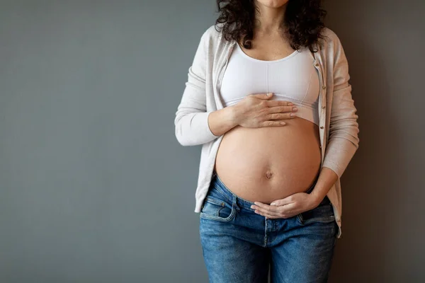 Pregnancy Concept. Cropped Shot Of Pregnant Young Lady Embracing Belly, Unrecognizable Expectant Woman Tenderly Touching Her Tummy While Standing Against Grey Wall Indoors, Copy Space