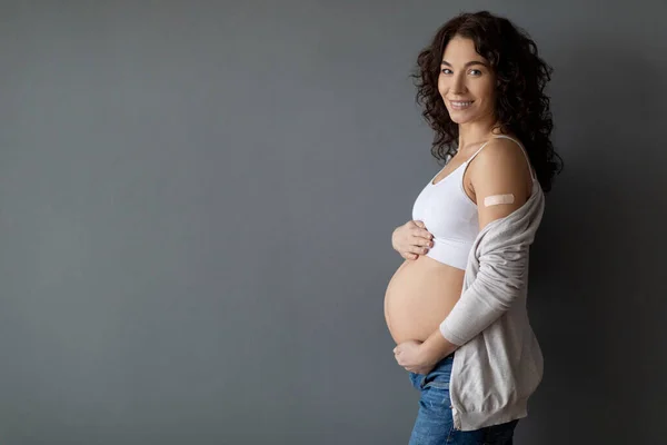 Pregnancy And Vaccination. Smiling Pregnant Woman Showing Arm With Adhesive Bandage After Vaccine Injection Shot, Vaccinated Expectant Female Standing Against Grey Wall Background, Copy Space