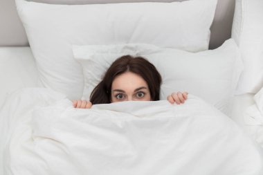 Beautiful big eyes and hands of playful young brunette woman fooling in bed at home, lady lying on pillows and hiding under blanket eiderdown, view above, copy space
