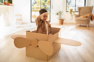 Happy small european kid plays in cardboard box helicopter, flies and fantasizes alone, looks at distance in children room interior. Dreams, art, entertainment with toys, fun and childhood at home