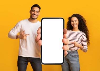 Nice Offer. Smiling Middle Eastern Couple Pointing At Big Blank Smartphone In Hands, Arab Man And Woman Demonstrating Copy Space For Mobile Advertisement, Standing On Yellow Background, Mockup