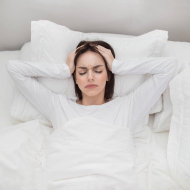 Top view of unhappy young brunette woman lying in bed at home, touching head with both hands, tired lady wake up with headache in the morning, suffering from chronic migraine, copy space