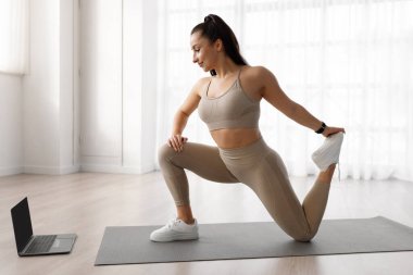 Side view of sporty beautiful millennial woman in sportswear training at home, stretching legs on fitness mat, looking at blank black computer screen, watching fitness video, copy space, mockup