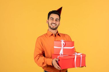 B-Day Party. Portrait Of Happy Handsome Arab Man Wearing Birthday Hat And Holding Wrapped Gift Boxes, Joyful Middle Eastern Guy Standing Isolated Over Yellow Studio Background, Copy Space
